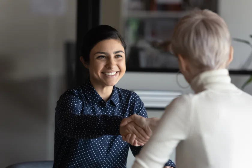 A professional woman smiling and shaking hands with a colleague in an office setting, emphasizing a positive and welcoming business interaction.