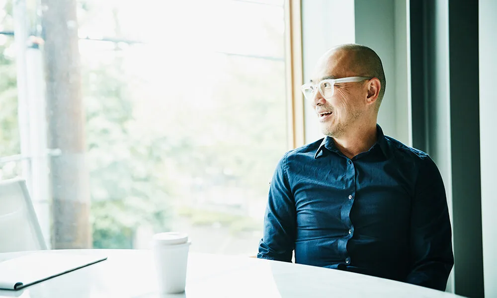 Business professional smiling while looking out a large office window, with natural light streaming in, conveying positivity and inspiration