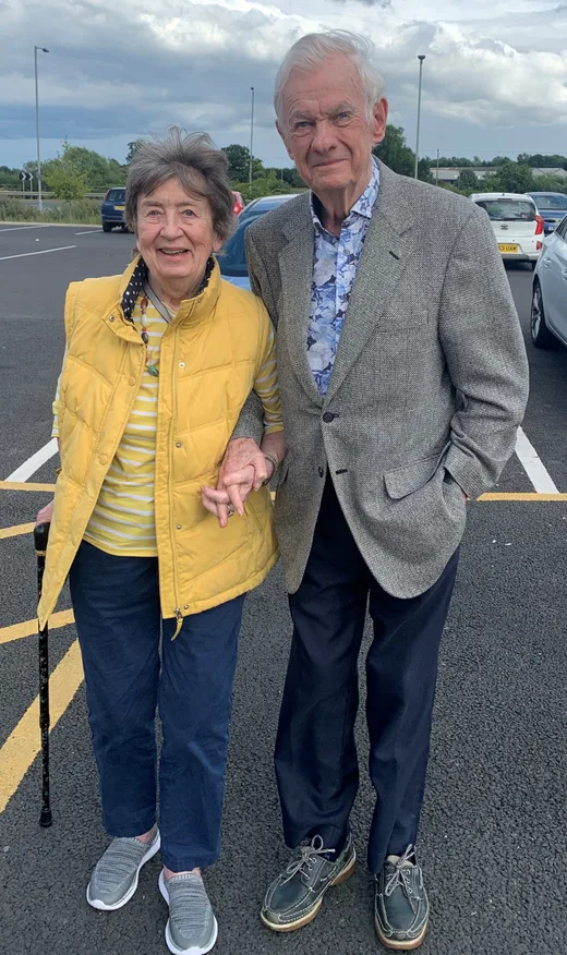 An elderly couple holding hands and smiling, with the woman in a yellow vest and the man in a grey blazer, standing in a parking lot