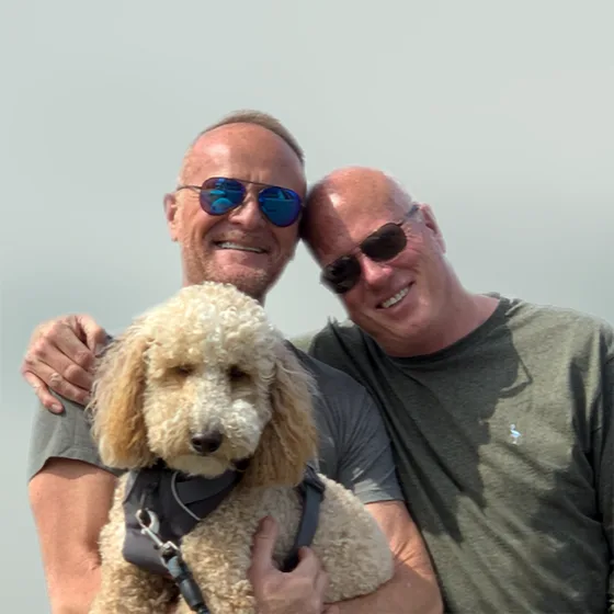 Two men smiling and posing outdoors with their pet poodle, symbolizing love and companionship during Pride Month