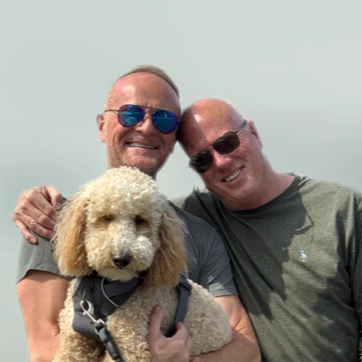 Two men smiling and posing outdoors with their pet poodle, symbolizing love and companionship during Pride Month