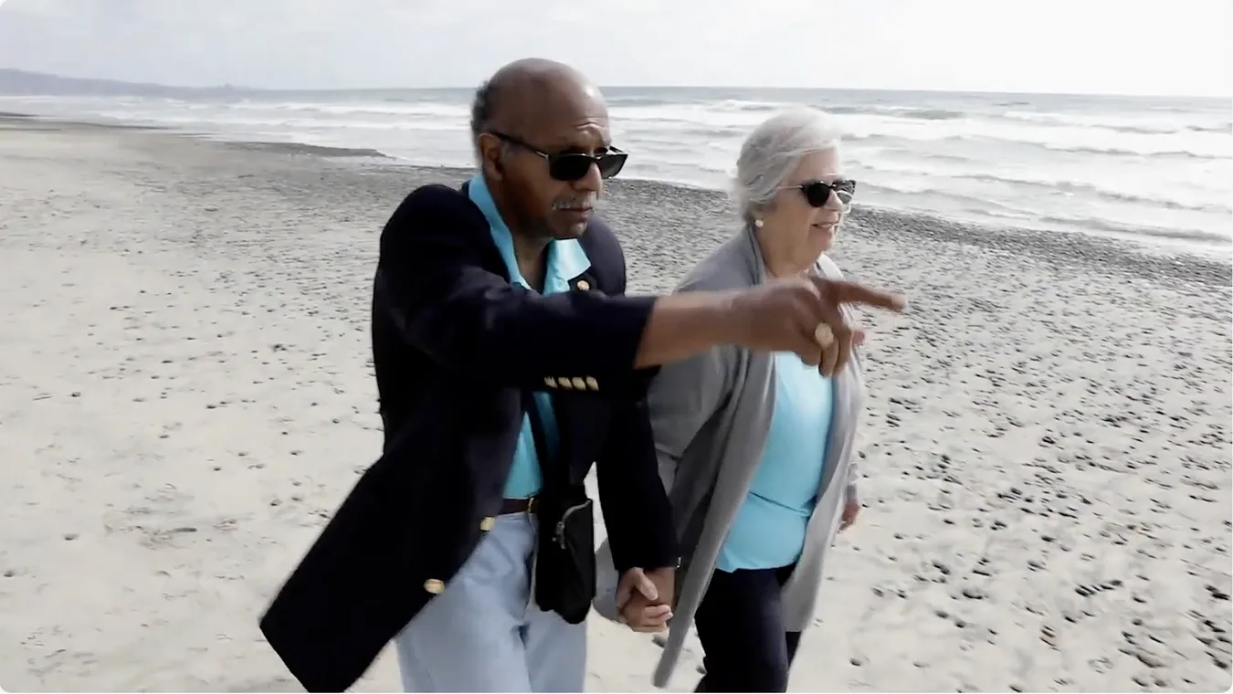 Elderly couple holding hands and walking along a beach, with one pointing towards the ocean, symbolizing companionship and enjoying life together