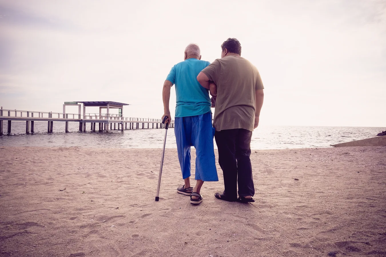 Two people walking on the beach, one supporting an elderly man using a cane while walking towards the ocean.