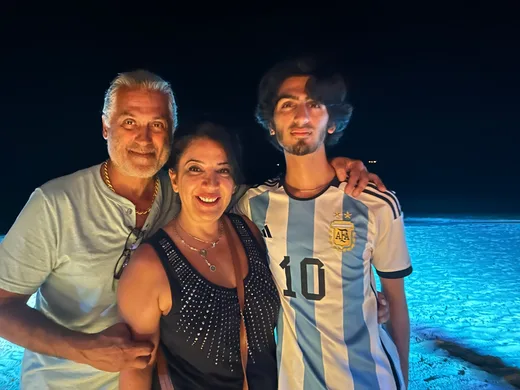 Three people smiling together at night on a beach, with the young man wearing an Argentina soccer jersey