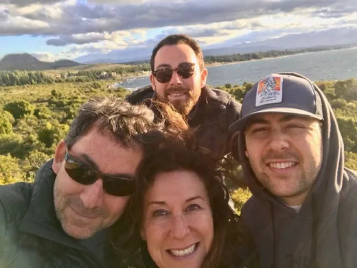 Family of four smiling outdoors with a scenic landscape of greenery, water, and mountains in the background