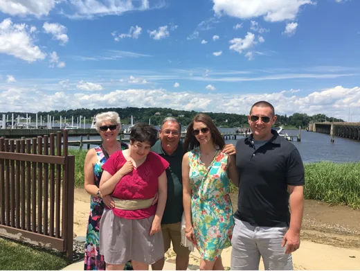 Family group photo by the waterfront on a sunny day, showing four adults and a young woman, all smiling together against a scenic background