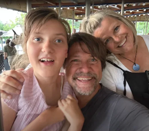 A family of three smiling, with the father and mother hugging their daughter with a disability, outdoors at an amusement park.