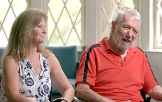 A female caregiver with an elderly man in a red shirt, seated and smiling in a bright room