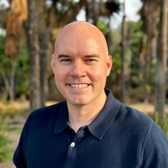 Portrait of a man with a bald head and a friendly smile, dressed in a navy polo shirt, standing outdoors with trees in the background