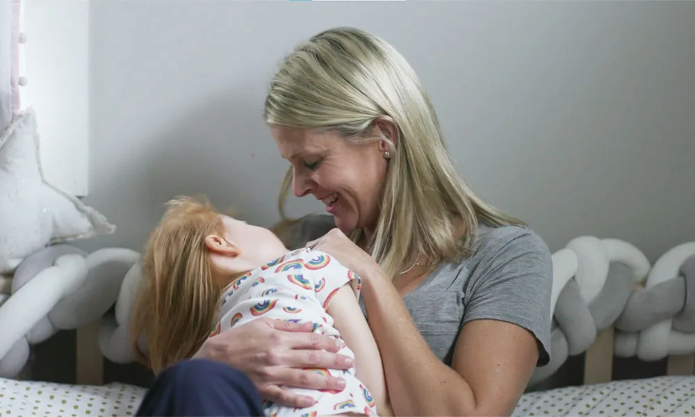 A mother embraces her young daughter in a bedroom, both sharing a tender moment