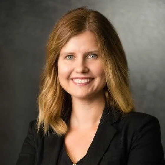 Professional headshot of a woman with blonde hair, smiling and dressed in a black blazer, standing against a dark background