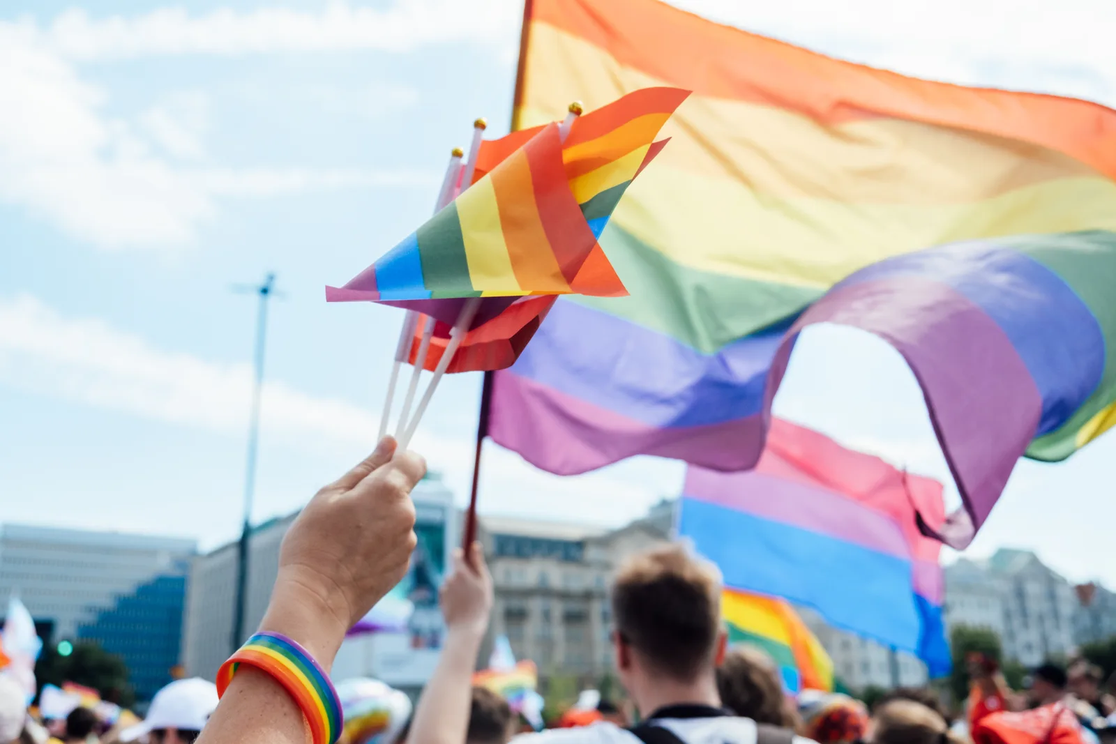 Close-up of hands holding multiple small Pride flags with a large rainbow flag in the background, symbolizing unity and celebration during a Pride Month event