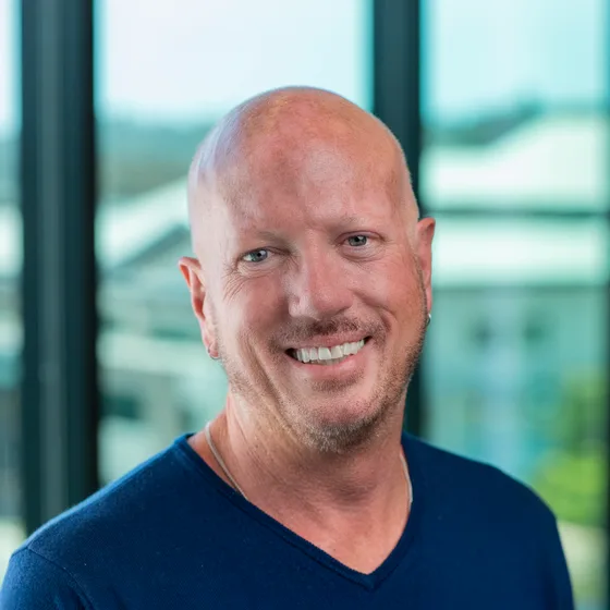 Professional headshot of a man with a shaved head, smiling and wearing a navy blue shirt, with a windowed background