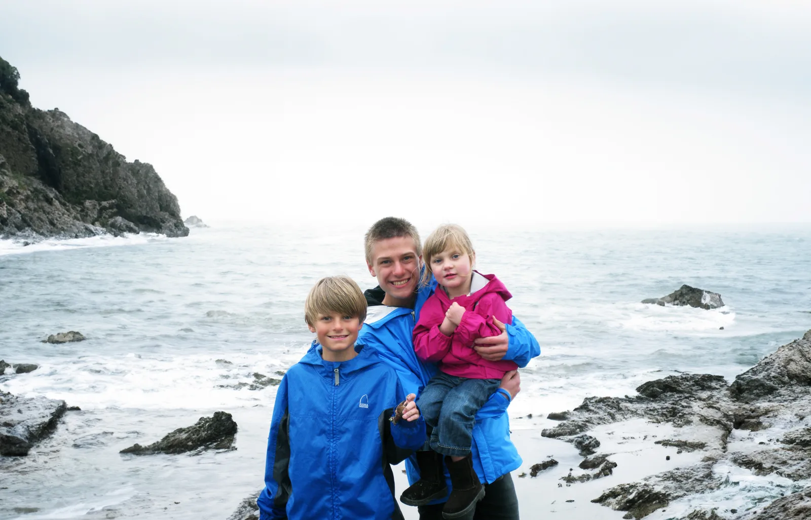 Three siblings, smiling and dressed warmly in colorful jackets, pose together on a rocky beach with the ocean in the background, symbolizing family resilience and connection