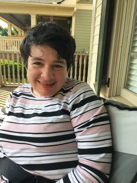 Young person smiling outdoors, dressed in a striped sweater, sitting on a porch with a peaceful and content expression