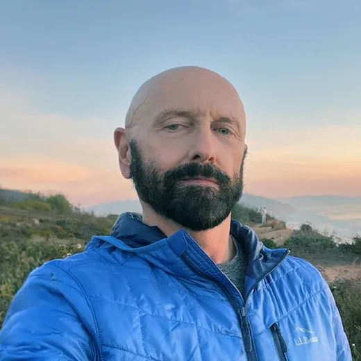 Portrait of a man with a bald head and a beard, wearing a blue jacket, standing outdoors with a sunset background