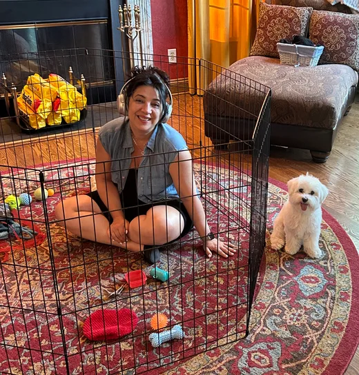 Woman sitting in a pet playpen with a happy white dog nearby, sharing a joyful moment in a cozy, warmly lit living room